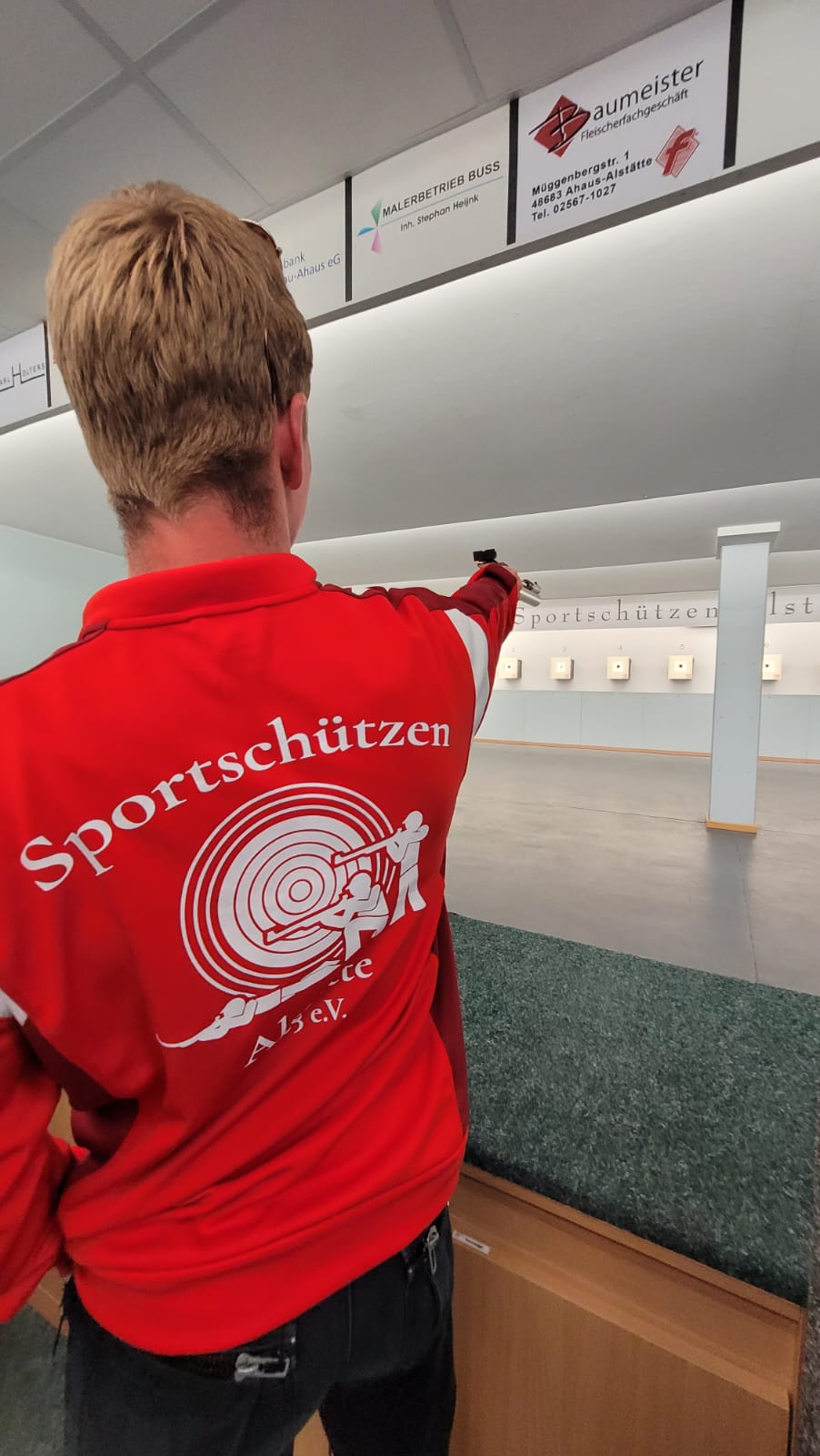 Junger Mann steht mit Rücken zur Kamera, wobei das Logo im Rücken im Fokus steht. Der Mann zielt mit einer Kurzwaffe auf eine Zielscheibe